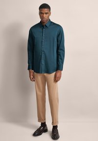 Dunkelblaugrünes Hemd mit langen Ärmeln, kombiniert mit hellbeigen Hosen und braunen Mokassins. Der Stoff sieht glatt und leicht glänzend aus.