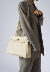 Cremfarbene Lederhandtasche mit strukturierter Form, goldener Hardware und abnehmbarem Schultergurt, gehalten von einer Person in einem grauen Anzug.