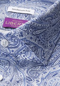 Col de chemise en tissu bleu à motifs cachemire avec un bouton blanc. Étiquettes indiquent "seidensticker" et "fabriqué avec un tissu Liberty." Surface texturée.