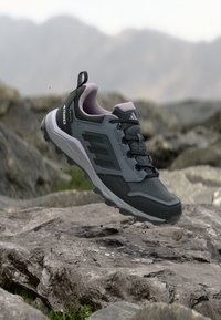 Tummanharmaa Adidas Terrex -vaelluskenkä ilmassa kivisen maaston yllä, taustalla epäselvä vuoristomaisema.