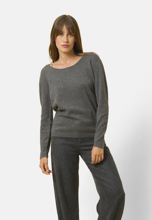 Maglione grigio a maglia con un ampio scollo e maniche lunghe, realizzato in materiale morbido. Abbinato a jeans scuri, in piedi su uno sfondo neutro.