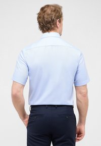 Man met een lichtblauwe kortafshirt met kraag, gestoken in een donkerblauwe broek, gezien vanuit de rug op een effen achtergrond.