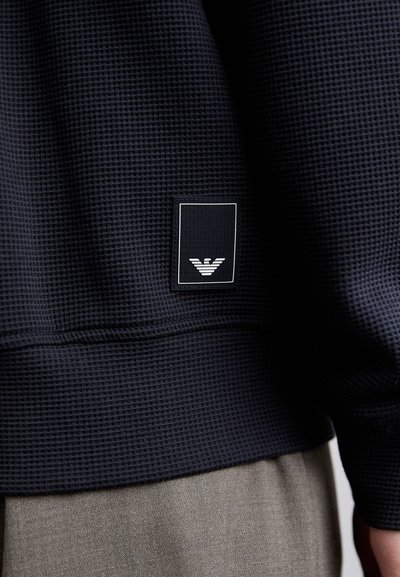 Tamsiai mėlynas džemperis su tekstūruota vaflinėmis raštomis ir baltu logotipo ženklu, vaizduojančiu stilizuotą erelį, kairėje apatinėje pusėje.