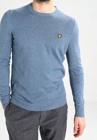 Maglione a maglia azzurro chiaro con collo rotondo, polsini e orlo a costine; presenta un piccolo logo giallo sul petto. Indossato con pantaloni grigi.