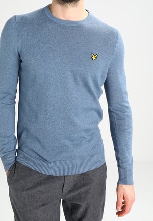 Pull en tricot bleu clair avec col rond, poignets et ourlet côtelés ; présente un petit logo jaune sur la poitrine. Porté avec un pantalon gris.
