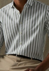 Stribet skjorte med knapper i hvidt og grønt stof med lange ærmer. Har en klassisk krave og en subtil tekstur. Kombineret med beige bukser.