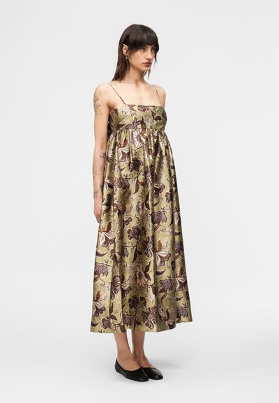 Γυναίκα που φοράει ένα μίντι φόρεμα με λουλούδια σε ελιά πράσινο χρώμα, χωρίς μανίκια, με λεπτές τιράντες και μαύρες φλατ, στέκεται μπροστά σε ένα απλό λευκό φόντο.