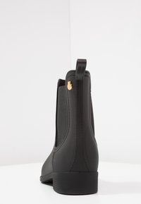 Czarny gumowy botek za kostkę z elastycznymi panelami bocznymi, z złotym akcentem logo, teksturowaną powierzchnią i niskim słupkowym obcasem. Gładkie wykończenie.