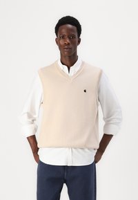 Μπεζ αμάνικο πουλόβερ με V-λαιμό, φορεμένο πάνω από λευκό πουκάμισο με κουμπιά. Διαθέτει ένα μικρό μαύρο λογότυπο στο στήθος.