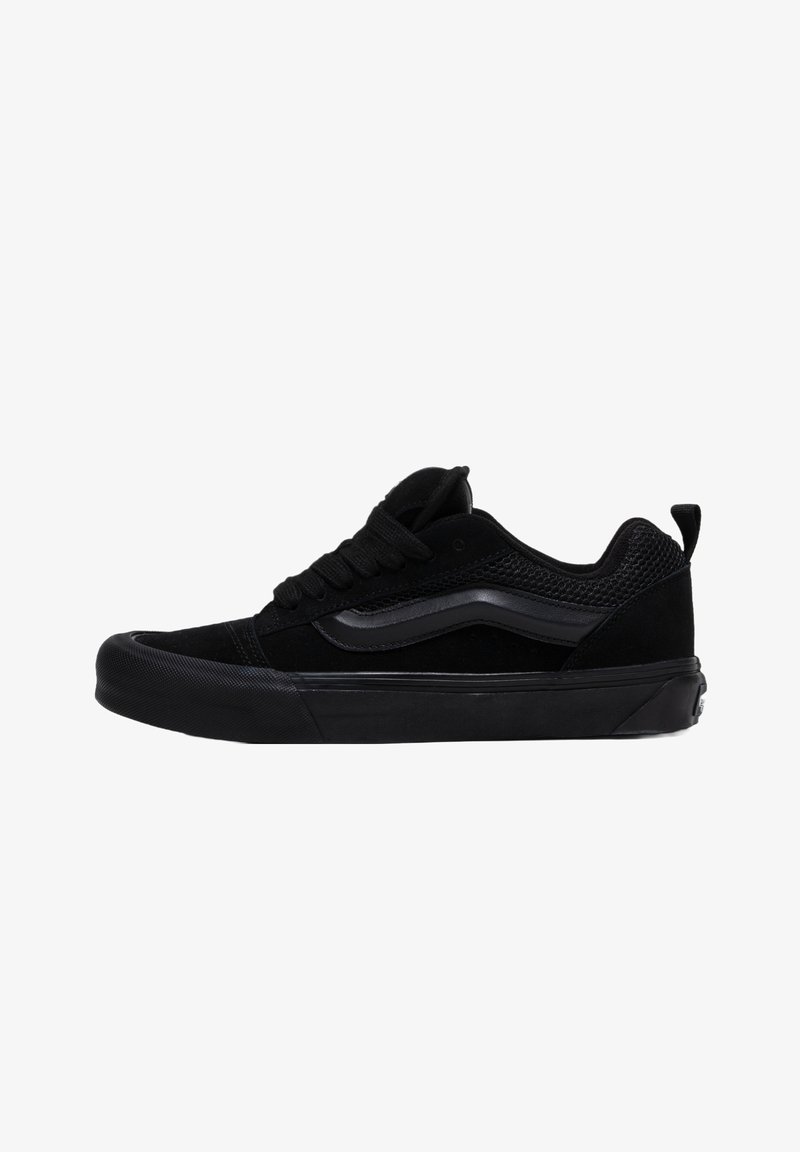 Chaussure de skate basse noire avec textures en mesh et daim, avec une bande latérale ondulée, une fermeture à lacets et une semelle en caoutchouc.