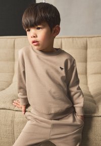 Beige Sweatshirt und passende Hose aus weichem Stoff. Pullover-Stil mit gerippten Bündchen und kleinem schwarzem Logo auf der Brust.