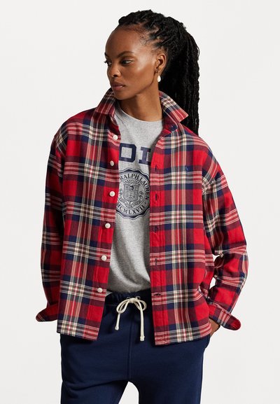 Chemise en flanelle à carreaux rouges et marine avec un design à boutons, portée sur un t-shirt graphique gris et un pantalon de survêtement marine. Présente un col.