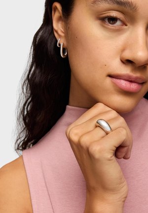 Una persona lleva pendientes de aro plateados y un anillo de plata liso y redondeado, que complementan una camiseta sin mangas ajustada de color rosa claro.