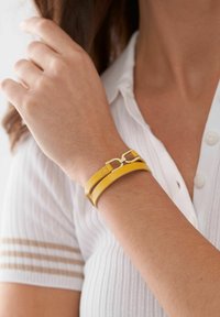 Yellow leather bracelet with gold-tone hardware, featuring two overlapping bands and a decorative clasp, worn on a wrist.