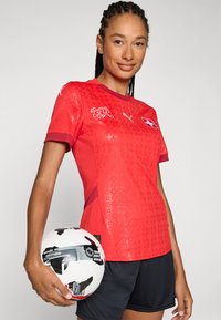 Red soccer jersey with short sleeves, textured pattern, and maroon accents. Black shorts worn, holding a black and white soccer ball with graphics.