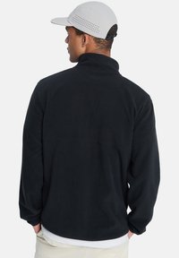Chaqueta de forro polar negra con cuello alto, mangas largas y una textura suave. Se lleva con una gorra gris claro que presenta orificios de ventilación.