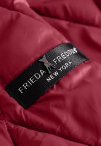 Zwart stoffen label met een klein hondenicoon en de tekst "FRIEDA FREDDIES NEW YORK" op een rode gewatteerde jasstof.
