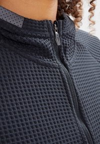 Veste zippée grise présentant un motif gaufré texturé, un col mandarin et une fermeture éclair. Tissu lisse avec une doublure intérieure à pois.