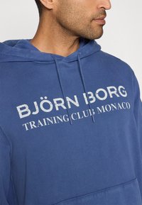 Kék kapucnis pulóver, pamut keverékből, a mellkasán kiemelkedő fehér felirattal: "BJÖRN BORG TRAINING CLUB MONACO".