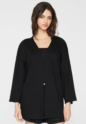 Frau mit langen dunklen Haaren, die eine schwarze, strukturierte offene Strickjacke und passende Shorts vor einem schlichten hellen Hintergrund trägt.