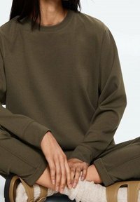 Sweat-shirt à manches longues vert olive en tissu texturé. Modèle assis, les mains croisées, portant des sandales beiges et des chaussettes de couleur crème.