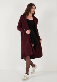 Cardigan burgundy cu dungi, cu mâneci lungi, deschis pe față, cu buzunare mari și accente de nasturi, purtat peste o rochie neagră și colanți.