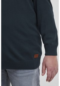 Tmavě zelený pletený svetr s žebrovanými rukávy a lemem. Má koženou nášivku na spodní části. Nosený s světle šedými džíny.