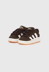 Hnědé semišové tenisky s bílými pruhy, šněrováním a bílou stélkou. Mají texturovanou oranžovou gumovou podrážku a logo.