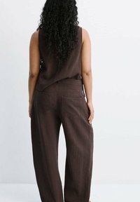 Femme aux longs cheveux bouclés portant un haut marron texturé sans manches et un pantalon large assorti, vue de dos sur un fond clair uni.