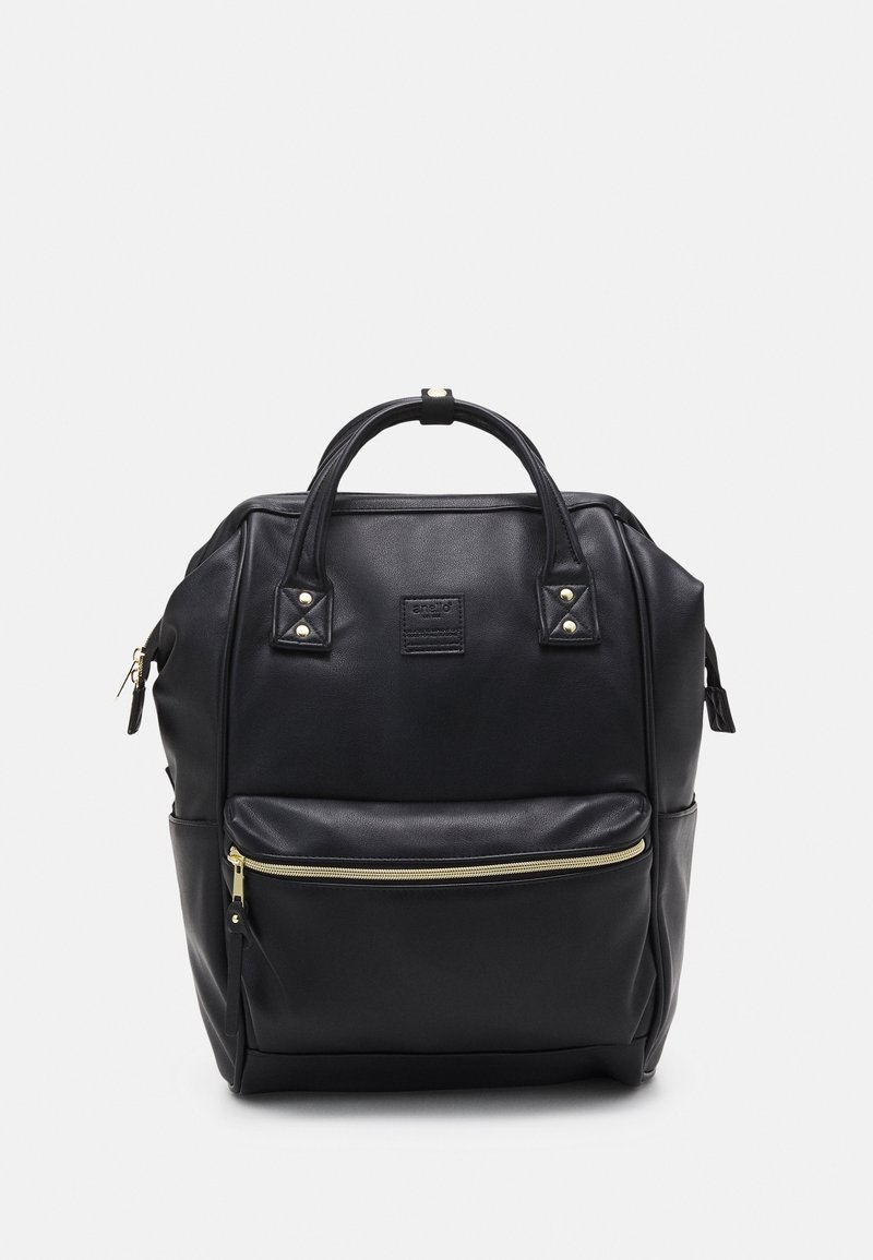 Sac à dos en cuir noir avec poignées supérieures, poche zippée à l'avant et fermeture éclair dorée sur fond gris clair.