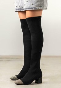 Botas de punto negras hasta la rodilla con un tacón grueso, que presentan una puntera metálica plateada en forma de punta y una textura suave.