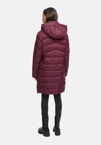 Mujer vista desde atrás que lleva un abrigo acolchado color burdeos hasta las rodillas con capucha, pantalones negros y botas negras de tobillo sobre un fondo blanco.