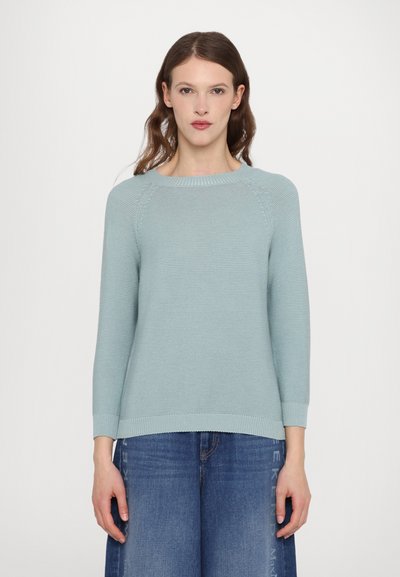 Lys blå strikket sweater med rund halsudskæring, lange raglanærmer og ribstrikket bund og manchetter, parret med brede denimbukser.