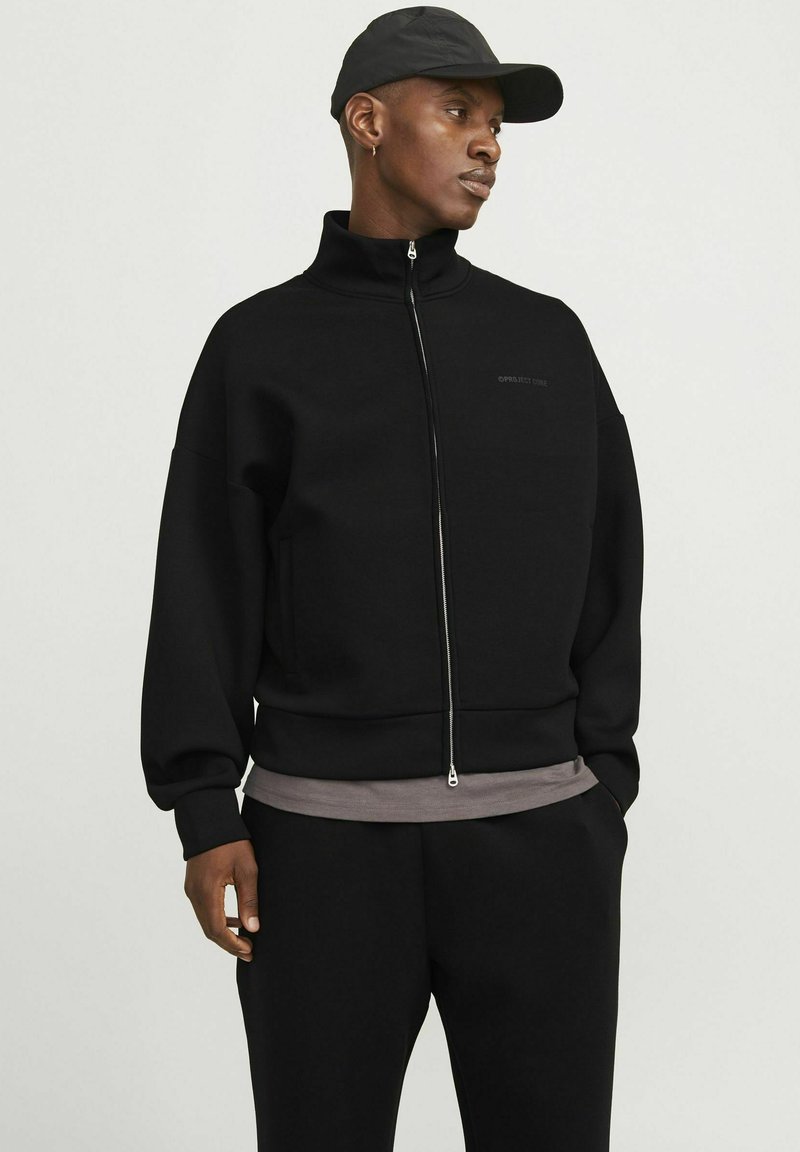 Sweatshirt noir zippé avec col haut, coupe oversize et poches avant. Porté sur un haut gris, associé à un pantalon noir et une casquette.
