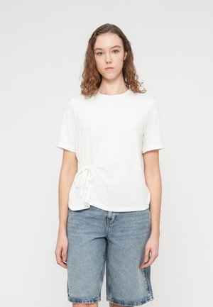 Maglietta bianca in cotone con maniche corte, caratterizzata da un dettaglio laterale attorcigliato. Abbinata a pantaloncini di jeans azzurri.