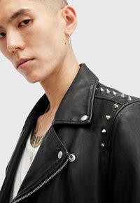 Jachetă din piele neagră de biker, cu studs pe umeri, accesorii din metal argintiu, închidere cu fermoar și finisaj texturat. Purtați peste o cămașă albă.