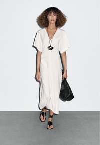 Witte jurk tot de knie met een V-hals en wijde mouwen, geaccessoreerd met een zwarte, hartvormige ketting en een geweven zwarte tas. Zwarte sandalen.