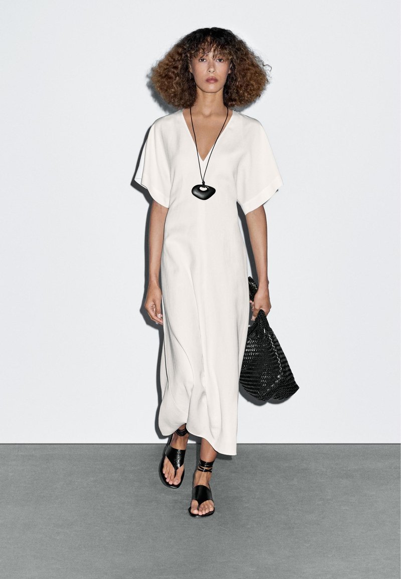 Witte jurk tot de knie met een V-hals en wijde mouwen, geaccessoreerd met een zwarte, hartvormige ketting en een geweven zwarte tas. Zwarte sandalen.