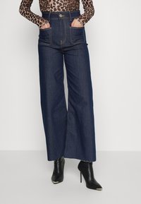 Pantalones de mezclilla oscuros de pierna ancha con una cintura alta, que presentan dos bolsillos frontales y costuras en contraste. Combinados con botines negros de punta.