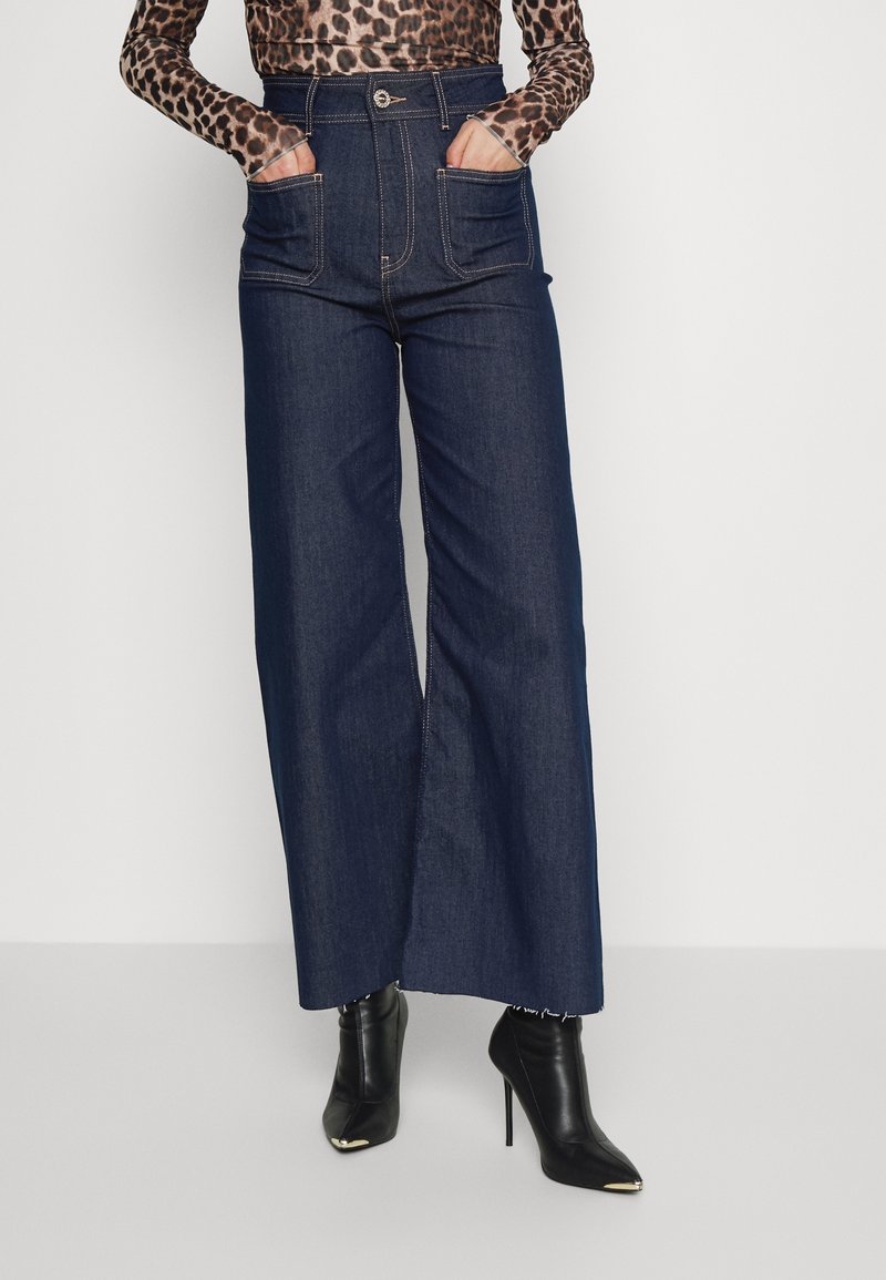 Pantalones de mezclilla oscuros de pierna ancha con una cintura alta, que presentan dos bolsillos frontales y costuras en contraste. Combinados con botines negros de punta.