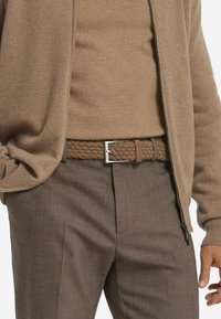 Geweven bruin riem met een zilveren gesp, gecombineerd met een tan kleurige lange mouw top en bruine textuur broek, die coördinerende neutrale tinten laat zien.