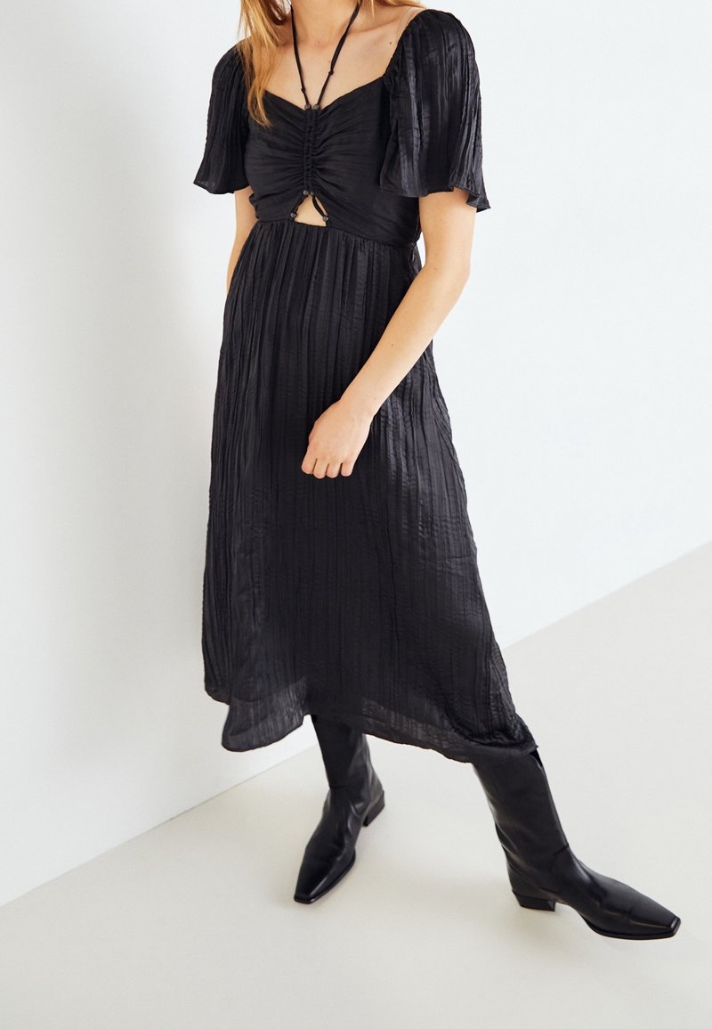 Vestido negro con textura, mangas cortas abullonadas y un corsé ajustado. Presenta un corte en forma de llave, falda fluida y se lleva con botas negras de punta.