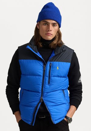 Blauwe puffer vest met een zwarte kraag en ritssluiting, voorzien van gestikte panelen en een logo op de borst, gedragen over een zwarte coltrui.
