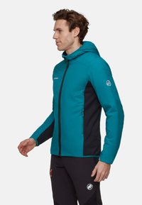 Homme de profil à gauche portant une veste à capuche teal avec des panneaux latéraux noirs et un pantalon noir avec un logo sur la cuisse.