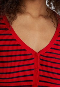Rood en marineblauwe gestreepte ribtop met een V-hals, voorzien van een knoopsluiting aan de voorkant en een structuurstof. De knopen zijn rood, in overeenstemming met de kraag.