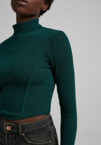 Maglione a coste aderente di un verde scuro, con colletto alto e maniche lunghe. Il tessuto testurizzato presenta linee verticali ed è tagliato corto.
