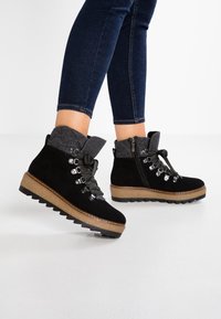 Botas de tobillo de ante negro con cuello de fieltro gris, parte delantera con cordones, ojales de metal y suela gruesa y texturizada con una plantilla intermedia marrón en contraste.