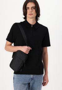 Svart polo-skjorte med korte ermer og glidelås i kragen, kombinert med lyseblå jeans. Modellen holder en liten svart crossover-veske.