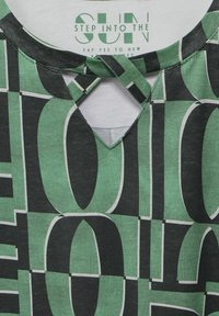 Top verde a fantasia con testo audace in bianco e nero, dotato di scollatura rotonda e un dettaglio di ritaglio unico sul retro. Tessuto morbido.