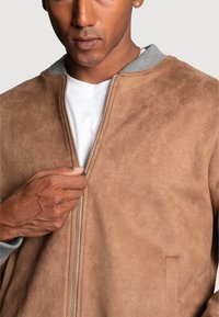 Veste en daim marron avec une fermeture éclair argentée et un col gris, présentant une texture lisse et des poches latérales, mettant en avant un design moderne.